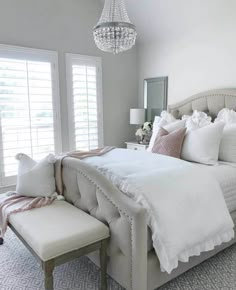 a white bed sitting in a bedroom next to a chandelier and two windows