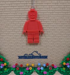 a red lego man hanging on a brick wall with flowers and plants around it in front of the sign