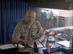 an older man sitting at a desk in front of a microphone and computer monitor with other equipment behind him