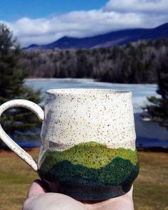 a hand holding a coffee mug in front of a lake with mountains and trees on it