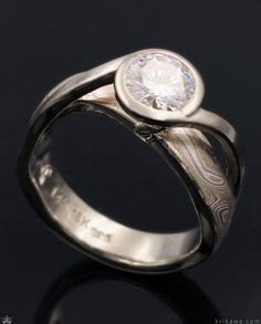 Mokume flows like a river around this band as it sweeps uninterrupted below the solitaire center stone. Metal bands emerge seamlessly from the band and form a bridge as they twist up and wrap the center stone in a bezel setting and suspend the solitaire over the mokume river below. Customized here in 14k white gold and Champagne Mokume Gane with a 0.70ct round moissanite solitaire. Twist Engagement Ring, Engagement Rings Twisted, Round Moissanite, Handcrafted Rings, A Bridge, Metal Bands, Solitaire Engagement Ring, Bezel Setting