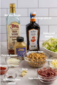 ingredients to make an italian salad laid out on a white counter top, including olives, lettuce, tomatoes, garlic, and wine vinegar