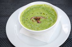 a white plate topped with a bowl filled with green soup and garnished with bacon