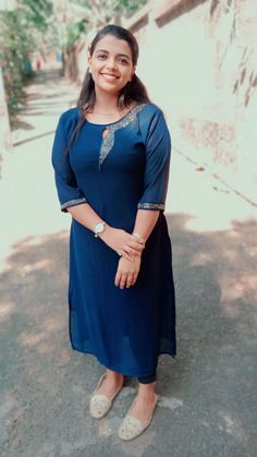 a woman in a blue dress is standing on the street smiling at the camera with her hands behind her back