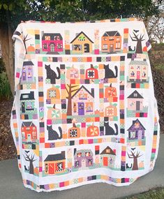 a quilt hanging on the side of a road with houses and trees in front of it