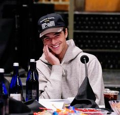 a man sitting at a table smiling and holding his hand to his face while wearing a hat