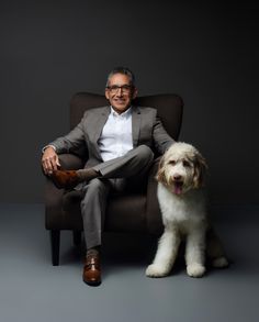 a man sitting in a chair next to a white dog with his legs crossed on the ground