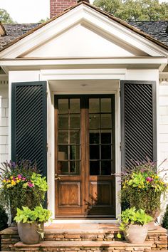 the front door to a white house with black shutters and two potted plants