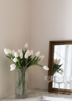 a vase filled with white flowers next to a mirror and wine glasses on a counter