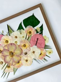 an arrangement of flowers is displayed in a wooden frame on the wall next to a plant