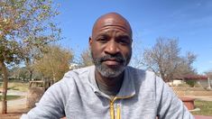 a man with a bald head sitting on a bench in a park and looking at the camera