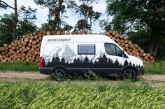 a van parked next to a pile of logs in a field with trees behind it