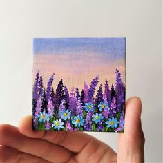 a hand holding up a small painting of purple and blue flowers