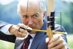 an older man in a suit and tie aiming with a bow at the target,