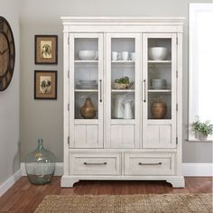 a white china cabinet with glass doors and drawers in a room next to a clock