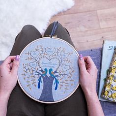 a woman holding up a cross - stitch tree with two birds on it next to a book