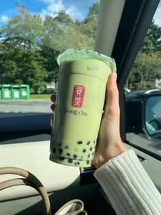 a person holding up a green drink in their hand while sitting in a car with the steering wheel behind them