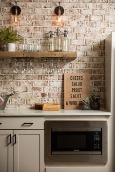 a kitchen with brick walls and white cabinets, an open shelf above the microwave is filled with wine glasses