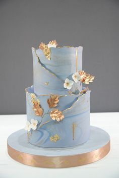 a three tiered blue cake with gold leaves and flowers on the top, sitting on a plate