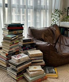 a pile of books sitting on the floor next to a brown leather chair in front of a window