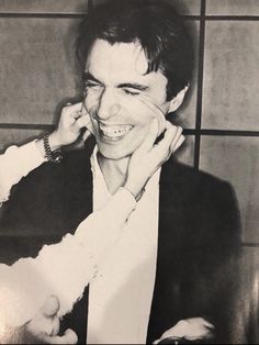 a black and white photo of a man smiling with his hand on his face while talking on the phone