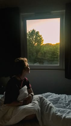 a little boy sitting on top of a bed next to a window with trees outside