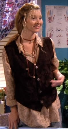 a woman with blonde hair wearing a brown vest and necklace standing in front of a plant