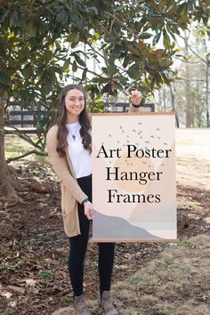 a woman standing next to a tree holding a sign that says art poster hanger frames