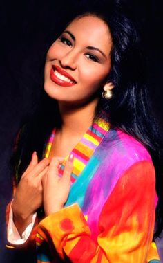 a woman with long black hair wearing a colorful shirt and earrings smiling at the camera