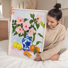 a woman sitting on a bed holding up a painting with pink flowers and lemons