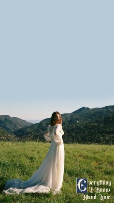 a woman in a long white dress standing on top of a lush green field with mountains in the background