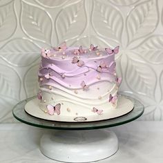 a white and pink cake with butterflies on it sitting on a glass plate in front of a wall