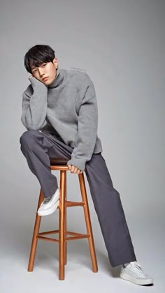 a man sitting on top of a wooden stool wearing a gray sweater and grey pants