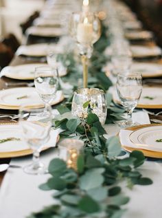 a long table is set with white plates and silverware, greenery and candlesticks