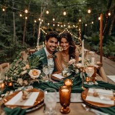 a man and woman sitting at a dinner table with candles in the middle, surrounded by greenery