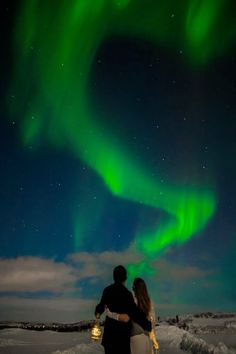 two people are standing in the snow under an auroral sky filled with green lights