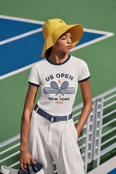 a young woman wearing a yellow hat and white pants stands in front of a tennis court