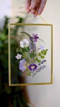 a hand holding a frame with flowers on it hanging from a wall next to a potted plant