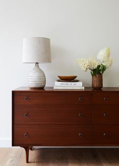 a white vase with flowers on top of a dresser next to a lamp and books