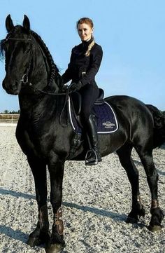 a woman riding on the back of a black horse in an enclosed area with gravel