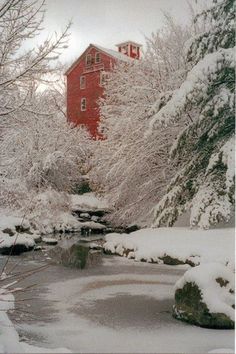 a red building surrounded by snow covered trees and water in the foreground is a small stream