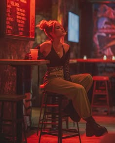 a woman sitting at a bar in front of a neon sign and drinking from a cup