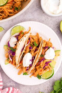 three shredded chicken tacos on a white plate with limes and sour cream