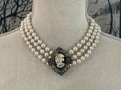 "This is the perfect sophisticated, gothic, edgy accessory for your Halloween look. This handmade necklace features a steampunk faux-cameo with a cream colored silhouette of a female skeleton with a gold bow in her hair on a black background and framed in gray filigree. The pendant sits in the center of three strands of faux-pearls.                Measurements:  -Length: 19 inches   -Pendant size: 2 inches x 1-1/2 inches ------------------------------------------------------------------------  Check out our \"BOO-tique\" for more fun Halloween-inspired pieces! For more lovely handmade, vintage and vintage-inspired items, visit our shop: www.etsy.com/shop/MadeleinesCollection" Elegant Black Jewelry For Cosplay, Elegant Halloween Jewelry For Costume Party, Elegant Jewelry For Halloween Costume Party, Steampunk Jewelry For Halloween Cosplay, Elegant Halloween Costume Jewelry, Elegant Jewelry For Cosplay, Elegant Adjustable Jewelry For Cosplay, Female Skeleton, Skull Cameo