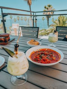 a table topped with plates of food and drinks next to the ocean on a sunny day