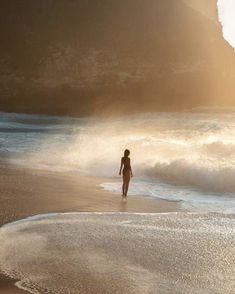 a woman walking on the beach in front of some water and mountains with waves coming up to her