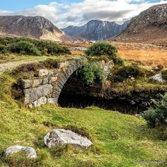 a stone bridge over a small stream in the middle of a field with mountains in the background