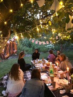 a group of people sitting around a table under a tree with lights strung over it