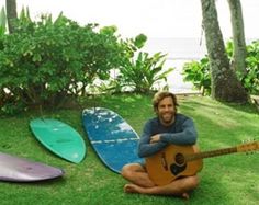 a man sitting on the grass with three surfboards behind him and holding a guitar