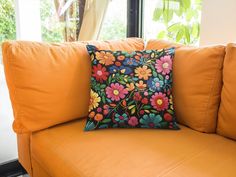 an orange couch with a colorful flowered pillow on it's back and window in the background